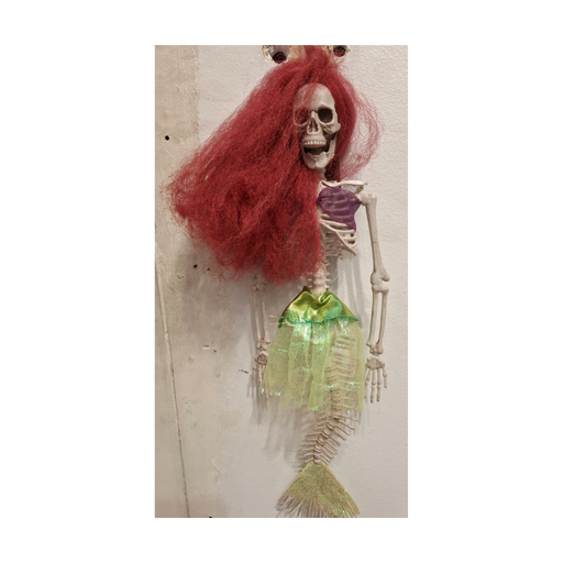 Esqueleto sirena colgante con pelo rojo, top morado y cola verde, colgado en un muro, foto real en tienda